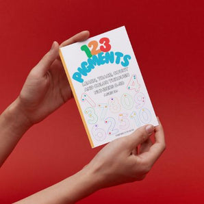 Hands holding Ermenrich Designs educational numbers coloring book against red background