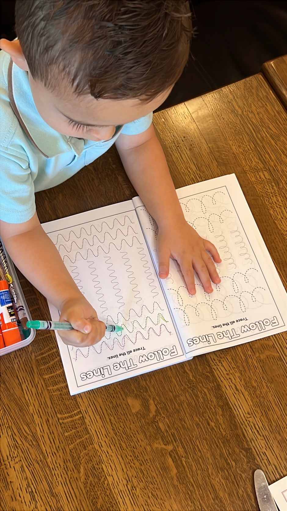 Child line tracing on activity coloring page in Preschool Essentials coloring book with crayons on a wooden table in a restaurant.