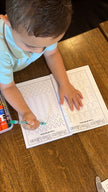 Child line tracing on activity coloring page in Preschool Essentials coloring book with crayons on a wooden table in a restaurant.