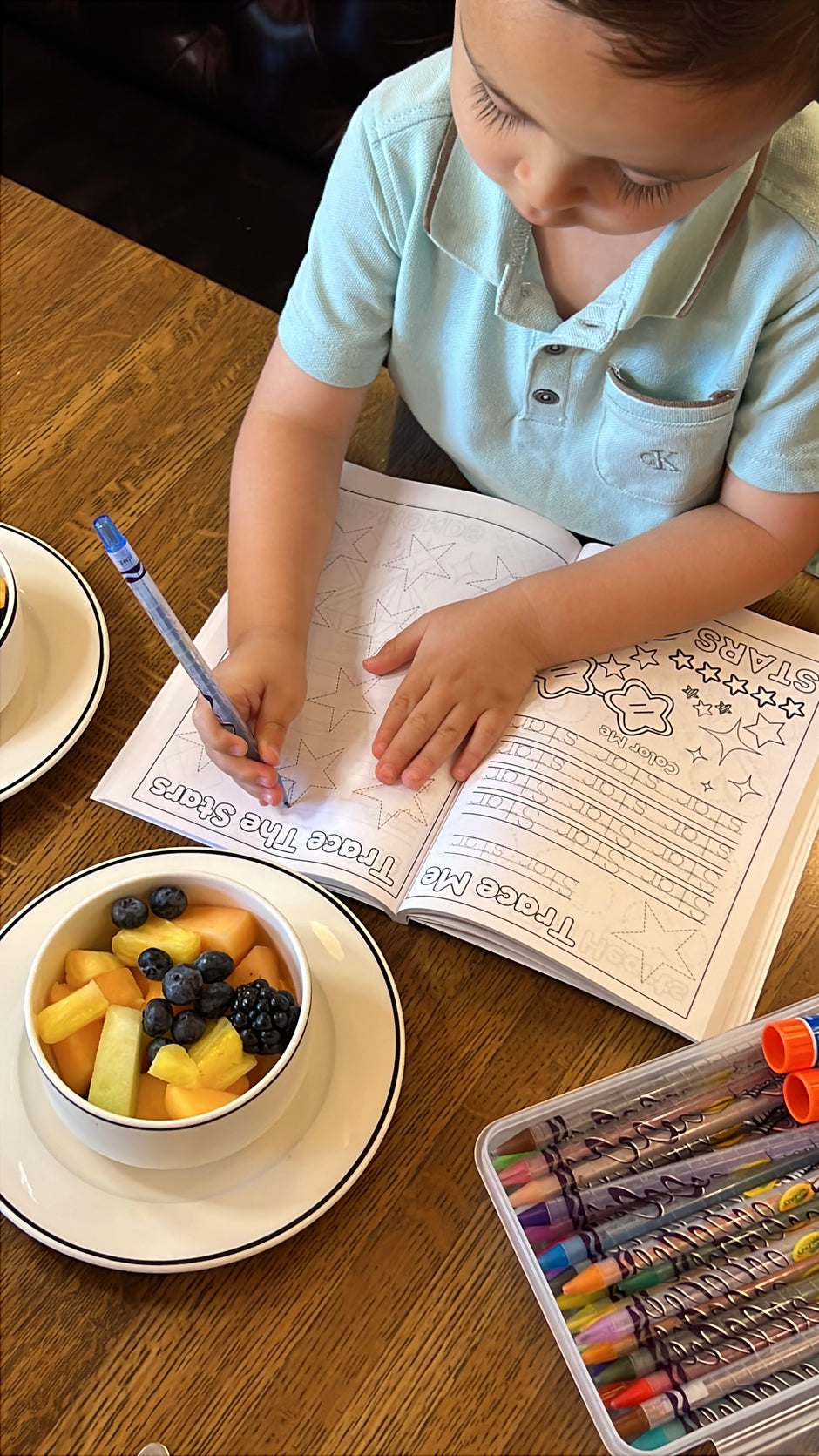 Child tracing stars in Preschool Essentials Trace Lines, Colors, Shapes and Numbers Coloring Book by Ermenrich Designs with a bowl of fruit and crayons on a table