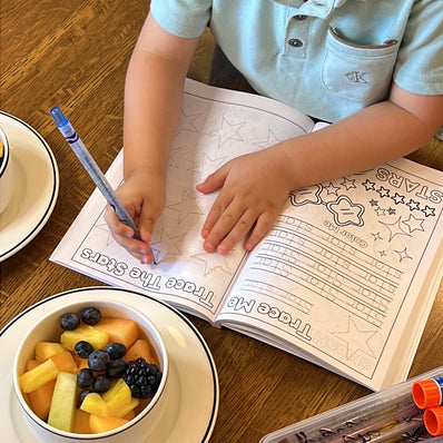 Child tracing stars in Preschool Essentials Trace Lines, Colors, Shapes and Numbers Coloring Book by Ermenrich Designs with a bowl of fruit and crayons on a table