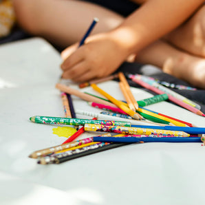 Colorful pencils on a white surface with a child's hand holding one, sitting on a light-colored floor.
